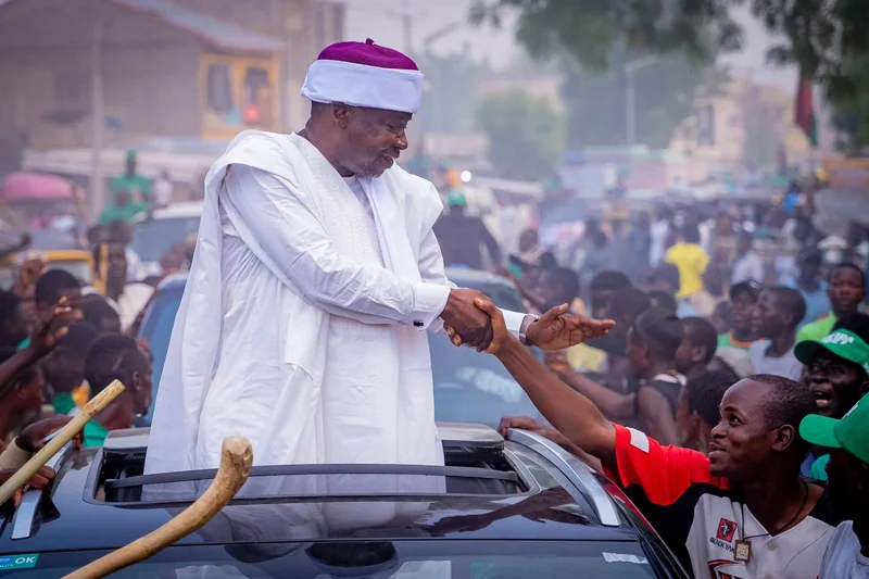 A man standing through a vehicle roof shakes hands with a person in the crowd.