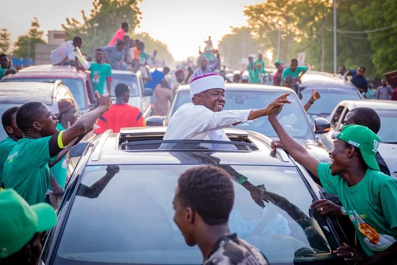 A man in white clothing greets people beside a vehicle on a busy street.