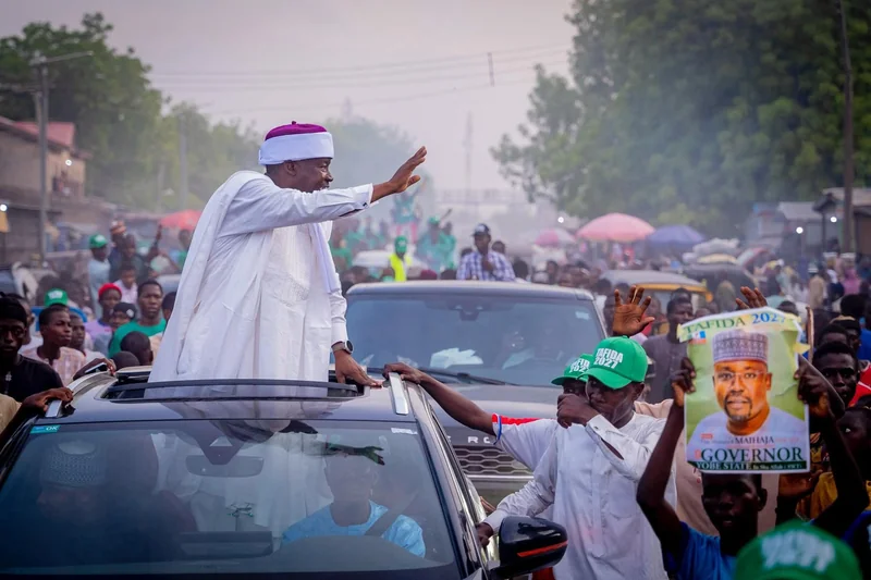 A man in white clothing waves from a vehicle surrounded by people.
